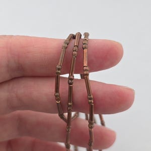 May include: A copper-toned necklace featuring a series of thin, cylindrical metal beads interspersed with small, round, textured spacer beads. The metallic chain has a warm, reddish-brown finish and a repeating pattern of long and short segments.