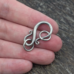 May include: A silver-toned metal toggle clasp featuring a curved, S-shaped bar with a textured, matte finish. The clasp includes a circular loop at one end and a decorative, spiraled hook design at the other, creating a secure interlocking mechanism.