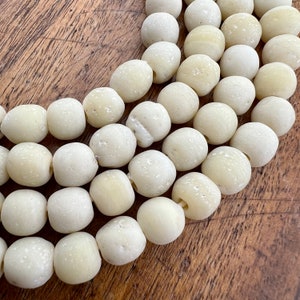 May include: A strand of off-white, round, smooth beads. The beads are arranged in a curved line, with the ends of the strand overlapping. The beads are all the same size and shape, and they have a slightly textured surface.