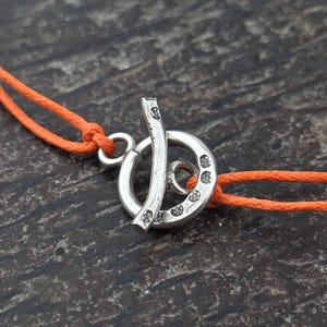 May include: A silver-toned toggle clasp featuring a circular ring and a T-bar component, both decorated with small circular indentations. The clasp is attached to two bright orange cords that extend outward from the central metal hardware.