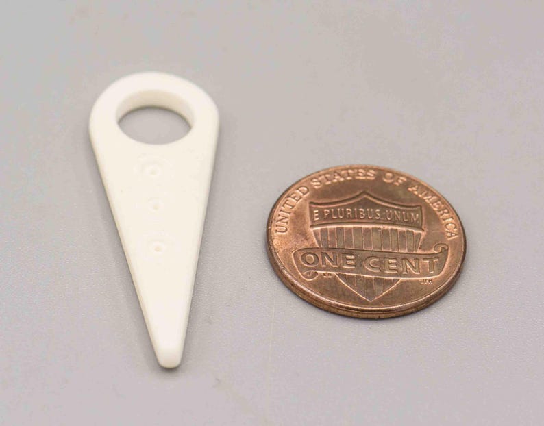 May include: A white plastic tool with a pointed tip and a small hole in the center. The tool is shaped like a triangle with a rounded top. A copper penny is in the background.
