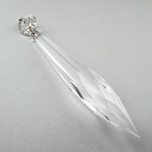 May include: A clear crystal prism with a faceted design and a small, round top. The prism is about 10 inches long and has a pointed tip.