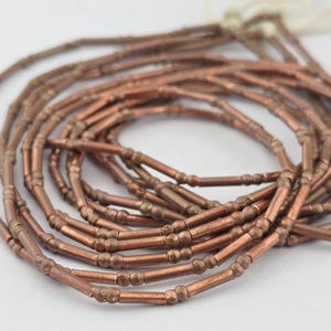 May include: A coiled strand of metallic copper-colored tube beads. Each bead features a long cylindrical center section flanked by two small spherical accents on either end. The beads are strung together on a thin white cord, creating a continuous loop.