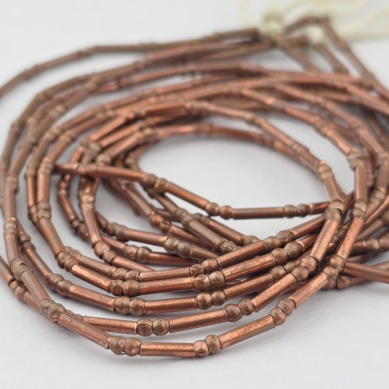 May include: A coiled strand of metallic copper-colored tube beads. Each bead features a long cylindrical center section flanked by two small spherical accents on either end. The beads are strung together on a thin white cord, creating a continuous loop.