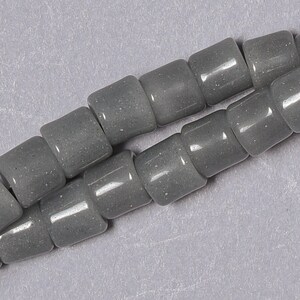 May include: A strand of gray glass beads with a speckled finish. The beads are cylindrical and have a slightly rounded shape.