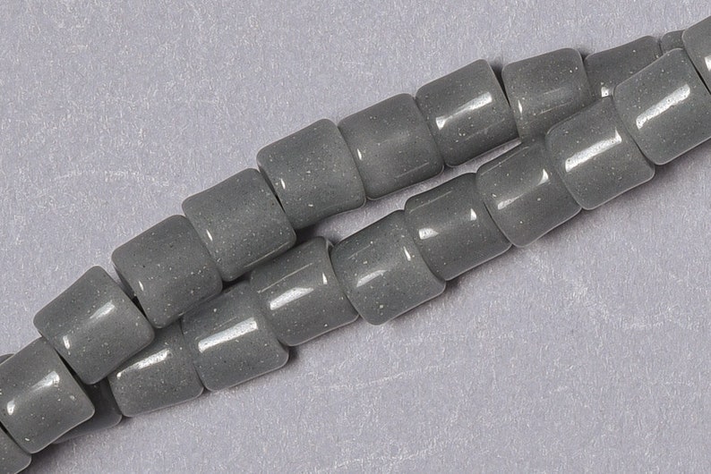 May include: A strand of gray glass beads with a speckled finish. The beads are cylindrical and have a slightly rounded shape.
