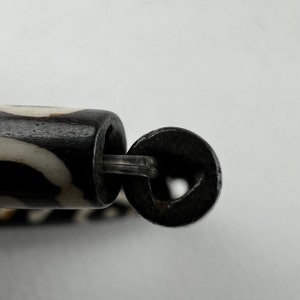 Puede incluir: Primer plano de una cuenta con estampado en blanco y negro con un anillo de metal oscuro unido por un conector de pl&aacute;stico transparente. La cuenta tiene forma cil&iacute;ndrica.