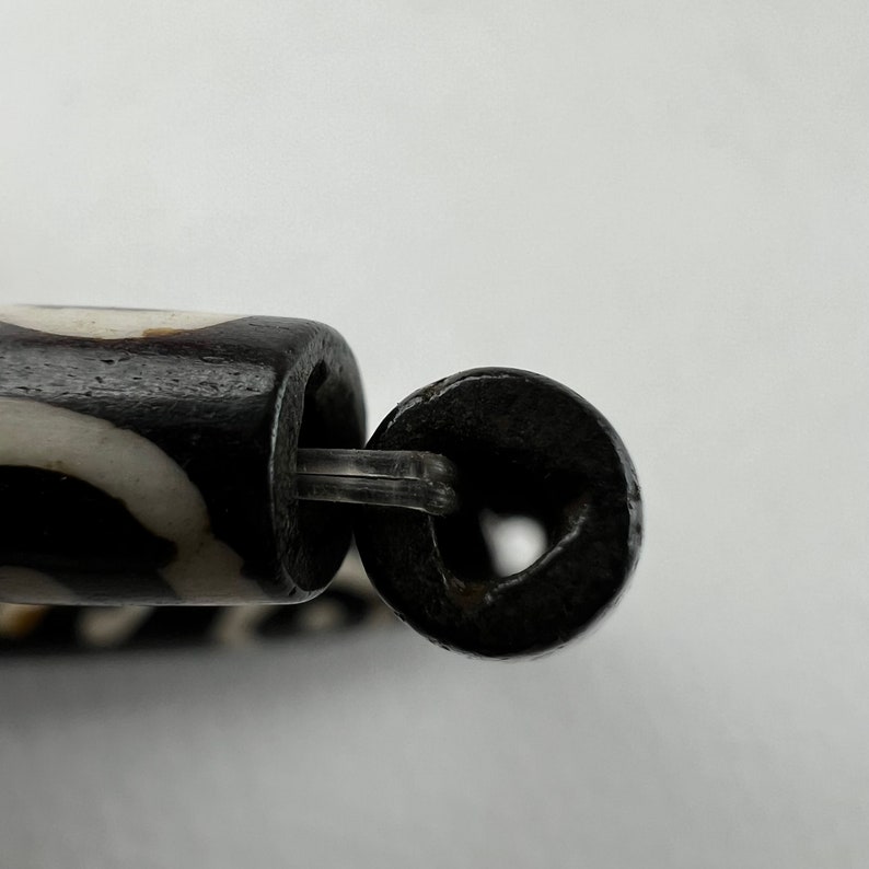 Puede incluir: Primer plano de una cuenta con estampado en blanco y negro con un anillo de metal oscuro unido por un conector de pl&aacute;stico transparente. La cuenta tiene forma cil&iacute;ndrica.