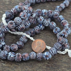 May include: A string of vintage glass beads featuring a distinctive striped design in blue, white, and maroon. The beads are strung on a white cord. A penny is next to the beads for size reference.