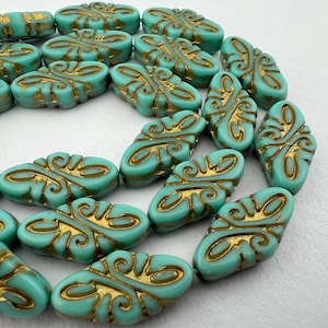 May include: A collection of turquoise-colored, oval-shaped beads with gold-colored, ornate designs. The beads have a slightly irregular shape and a glossy finish. These beads are suitable for jewelry making.