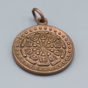 May include: A circular bronze pendant with a detailed, intricate design. The pendant features a repeating pattern of geometric shapes and a central point.