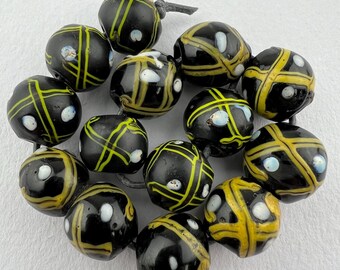 Pode incluir: Um fio de contas de vidro preto com detalhes amarelos e brancos. As contas têm um padrão de cruz e são enfiadas em um cordão.