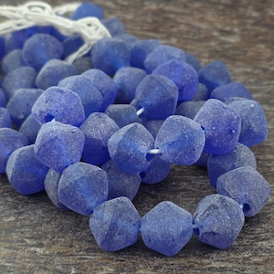 May include: Close-up of a collection of faceted, cobalt blue glass beads. The beads are strung on a white cord, with a few loose beads scattered around. The beads have a frosted appearance and a geometric shape. The background is a dark, textured surface.