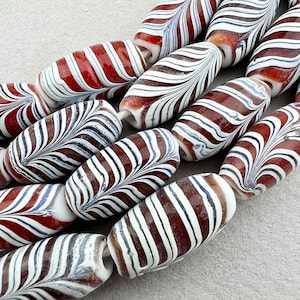 May include: A collection of red and white striped glass beads with a marbled effect. The beads are oval shaped and have a glossy finish.
