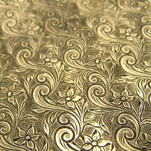 May include: A close-up of a gold-colored surface with an embossed floral pattern. The pattern features intricate swirls and leaves, creating a delicate and elegant design.