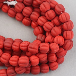 May include: A strand of red, matte-finish beads. Each bead is a rounded rectangle with carved horizontal lines, resembling small pumpkins. The beads are strung together on a white string, creating a continuous loop. The background is a neutral gray.