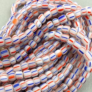 Peut inclure: Un brin de perles de verre blanches avec des rayures rouges et bleues. Les perles sont petites et rondes, et elles sont enfilées sur un fil transparent.