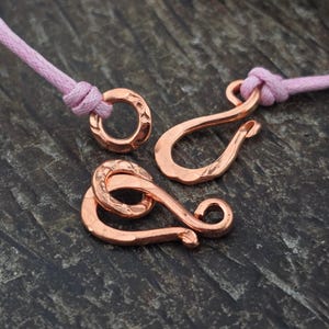 May include: A set of copper-colored metal jewelry clasps featuring a hammered texture. The set includes a circular ring and an S-shaped hook, both attached to light pink cord segments. The metal components have a rustic, hand-forged appearance.