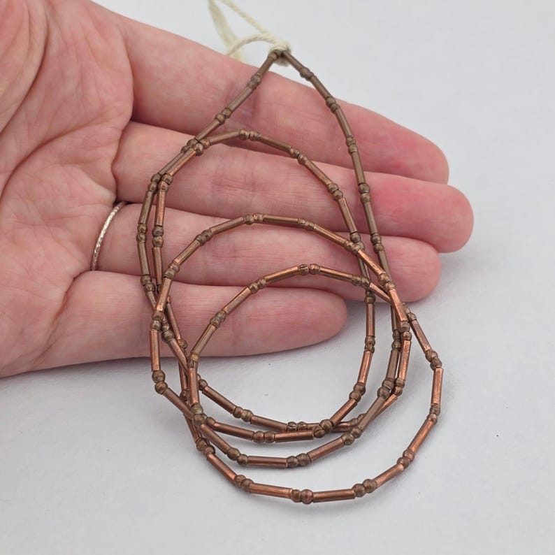 May include: A strand of copper-colored tubular beads with small spherical accents at regular intervals. The beads are strung on a thin white cord, forming a series of concentric loops that showcase the metallic finish and the repeating pattern.