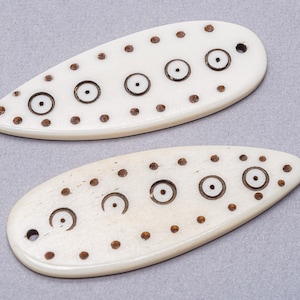May include: Two off-white, teardrop-shaped pendants with a series of dark brown dots and circles. Each pendant has a small hole at the pointed end. The pendants are displayed on a gray background.