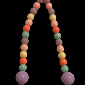 May include: A colorful beaded necklace with a heart-shaped pendant. The necklace features a variety of pastel-colored beads, including pink, orange, yellow, green, and purple. The pendant is a textured, light purple heart.