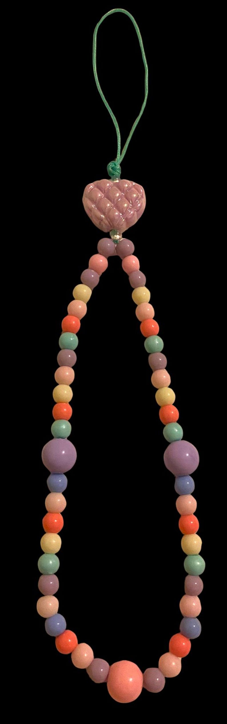 May include: A colorful beaded necklace with a heart-shaped pendant. The necklace features a variety of pastel-colored beads, including pink, orange, yellow, green, and purple. The pendant is a textured, light purple heart.