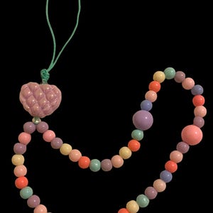 May include: A colorful beaded necklace with a quilted heart pendant. The necklace features a variety of pastel-colored beads, including pink, orange, yellow, green, and purple. The heart pendant is a light pink color and is attached to a green cord.