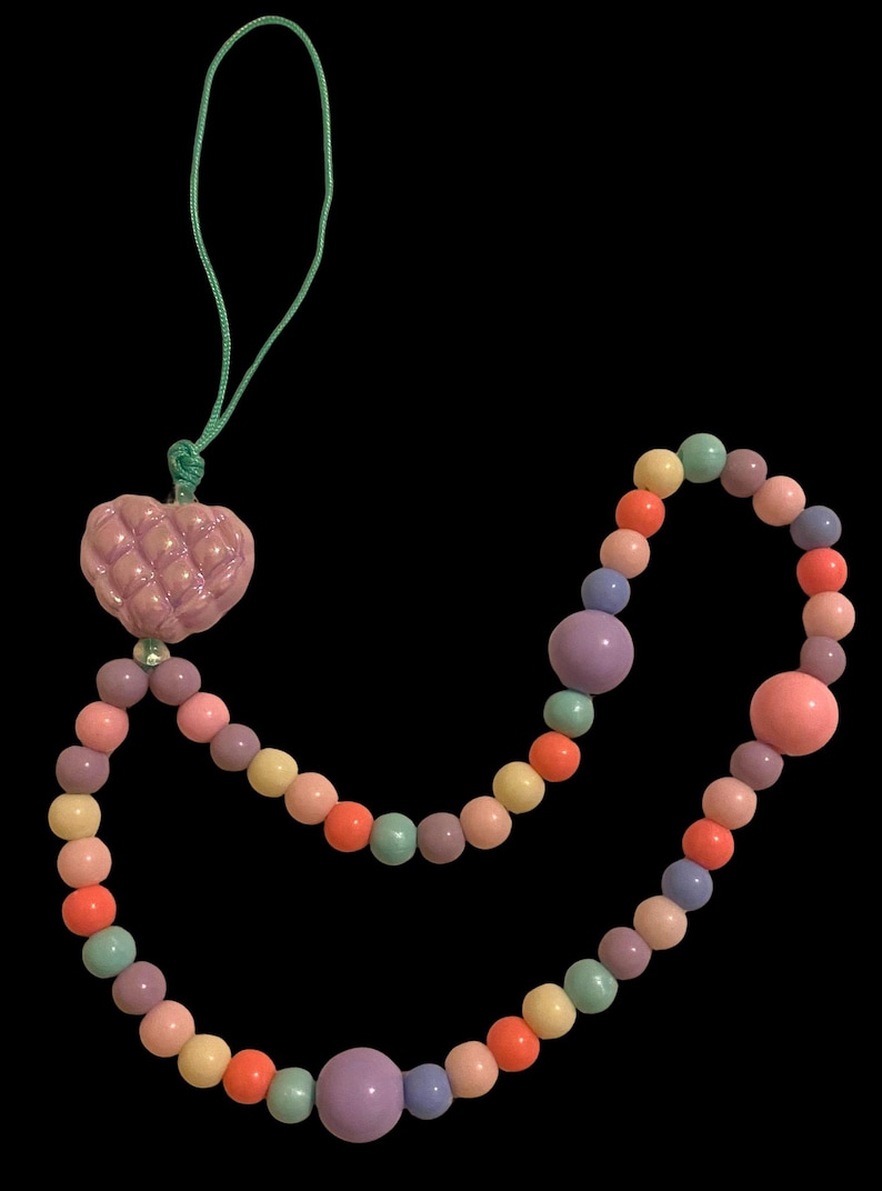 May include: A colorful beaded necklace with a quilted heart pendant. The necklace features a variety of pastel-colored beads, including pink, orange, yellow, green, and purple. The heart pendant is a light pink color and is attached to a green cord.