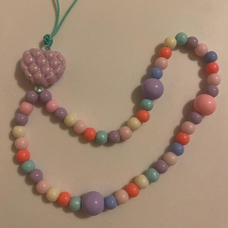 May include: A pastel-colored beaded necklace with a heart-shaped pendant. The necklace features a variety of round beads in shades of pink, purple, orange, yellow, and blue. The heart pendant is a light purple color and has a textured surface.
