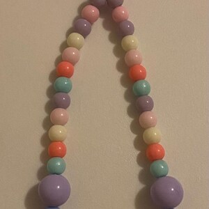 May include: A pastel-colored beaded necklace with a heart-shaped pendant. The necklace features a variety of round beads in shades of pink, purple, orange, yellow, and blue. The pendant is a light purple heart.