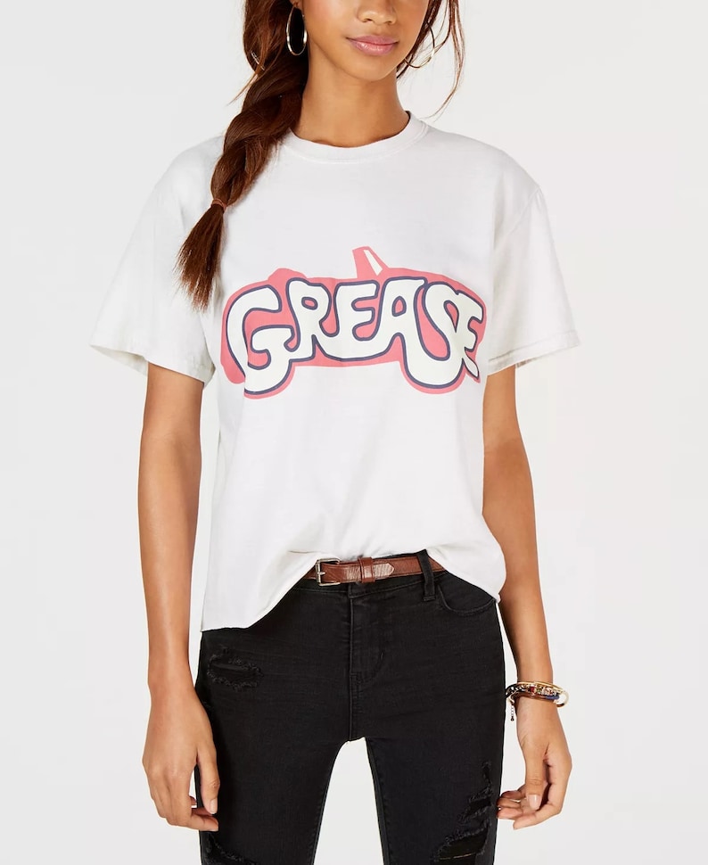 Pu&ograve; includere: Maglietta bianca con un logo rosa "Grease" che presenta la silhouette di un'auto.