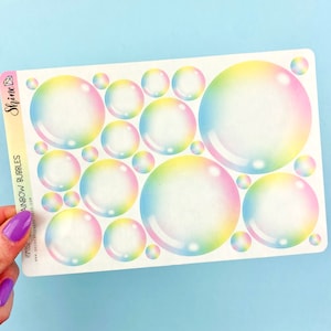 Puede incluir: Una hoja de pegatinas de burbujas arcoíris. Las pegatinas son transparentes con un degradado arcoíris. La hoja es blanca con el texto "Rainbow Bubbles" y "Shine Sticker Studio" impreso en ella.