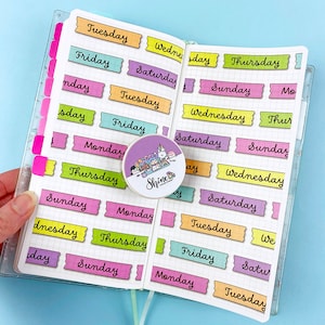 May include: A set of colourful washi tape stickers with the days of the week written on them. The stickers are in a variety of colours, including pink, yellow, green, and blue. The stickers are arranged in a grid pattern on a white background.