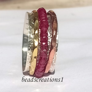 May include: A multi-band ring featuring a central band of small, faceted red gemstones. The ring has bands of silver, gold, and copper-toned metal with textured and hammered finishes. The text "beadscreations1" is at the bottom.