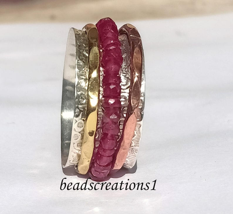 K&ouml;nnte beinhalten: Ein mehrreihiger Ring mit einem zentralen Band aus kleinen, facettierten roten Edelsteinen. Der Ring hat B&auml;nder aus Silber, Gold und kupferfarbenem Metall mit strukturierten Oberfl&auml;chen. Der Text "beadscreations1" befindet sich unten.