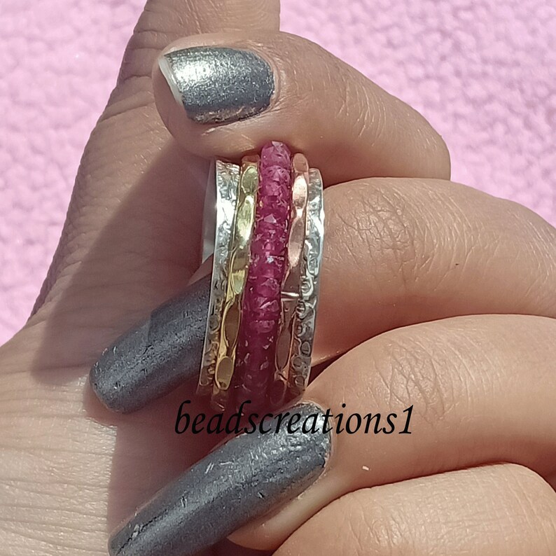 K&ouml;nnte beinhalten: Ein silberner, goldener und kupferfarbener Spinner-Ring mit einem zentralen Band aus kleinen, grob geschnittenen, rubinroten Steinen. Der Ring hat ein strukturiertes Design und der Text "beadscreations1" ist sichtbar.