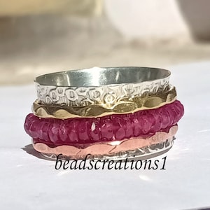 K&ouml;nnte beinhalten: Ein Stapel von Spinner-Ringen in Silber, Gold, Ros&eacute;gold und einem tiefen Magenta. Die Ringe haben strukturierte und glatte B&auml;nder. Der Text "beadscreations1" befindet sich unten rechts im Bild.