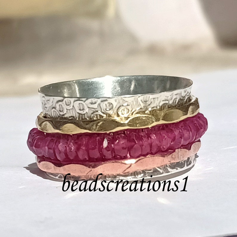 K&ouml;nnte beinhalten: Ein Stapel von Spinner-Ringen in Silber, Gold, Ros&eacute;gold und einem tiefen Magenta. Die Ringe haben strukturierte und glatte B&auml;nder. Der Text "beadscreations1" befindet sich unten rechts im Bild.