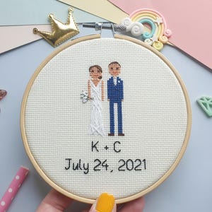 Puede incluir: Un bastidor de bordado de punto de cruz con una escena de boda que muestra a una novia y un novio. La novia lleva un vestido blanco y sostiene un ramo, mientras que el novio lleva un traje azul. El texto "K + C July 24, 2021" est&aacute; bordado debajo.