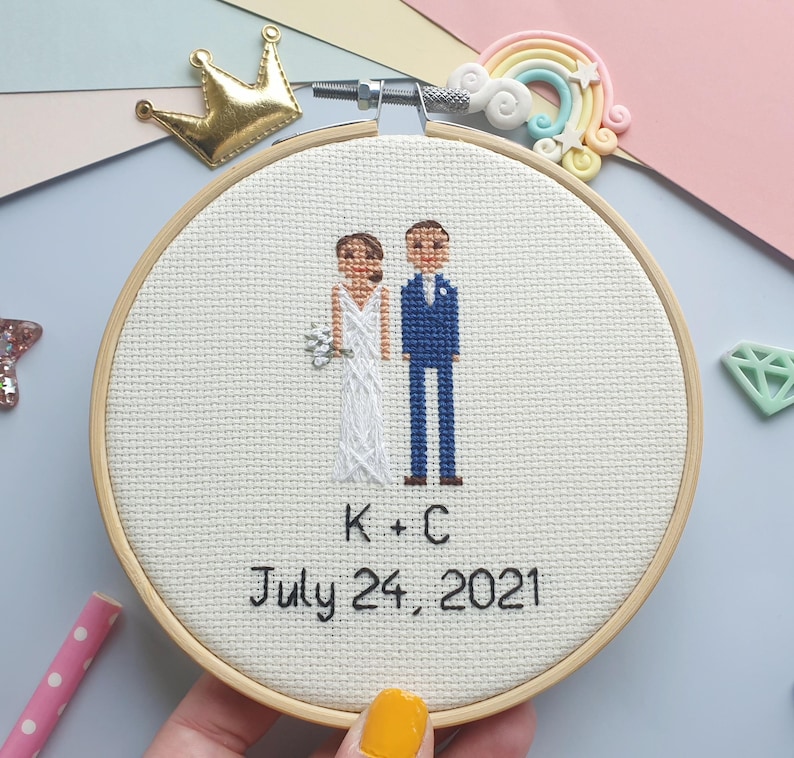 Puede incluir: Un bastidor de bordado de punto de cruz con una escena de boda que muestra a una novia y un novio. La novia lleva un vestido blanco y sostiene un ramo, mientras que el novio lleva un traje azul. El texto "K + C July 24, 2021" est&aacute; bordado debajo.
