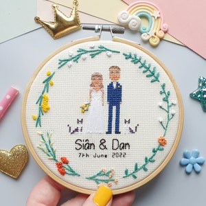 A cross stitch portrait framed in a wooden embroidery hoop with 2 stitched people in wedding attire with their 2 cats on either side of them. The bride is holding a yellow & orange bouquet that matches the floral border around the outside of the hoop