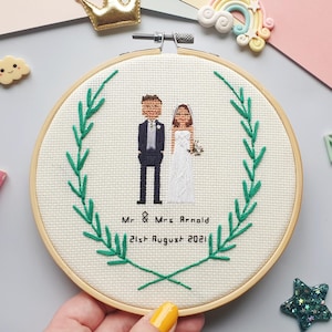 Puede incluir: Un bordado de punto de cruz circular en un aro de madera, que representa a una pareja de bodas dentro de una corona de hojas verdes. El texto "Mr & Mrs Arnold 21st August 2021" est&aacute; bordado debajo. El fondo es de tela beige claro.