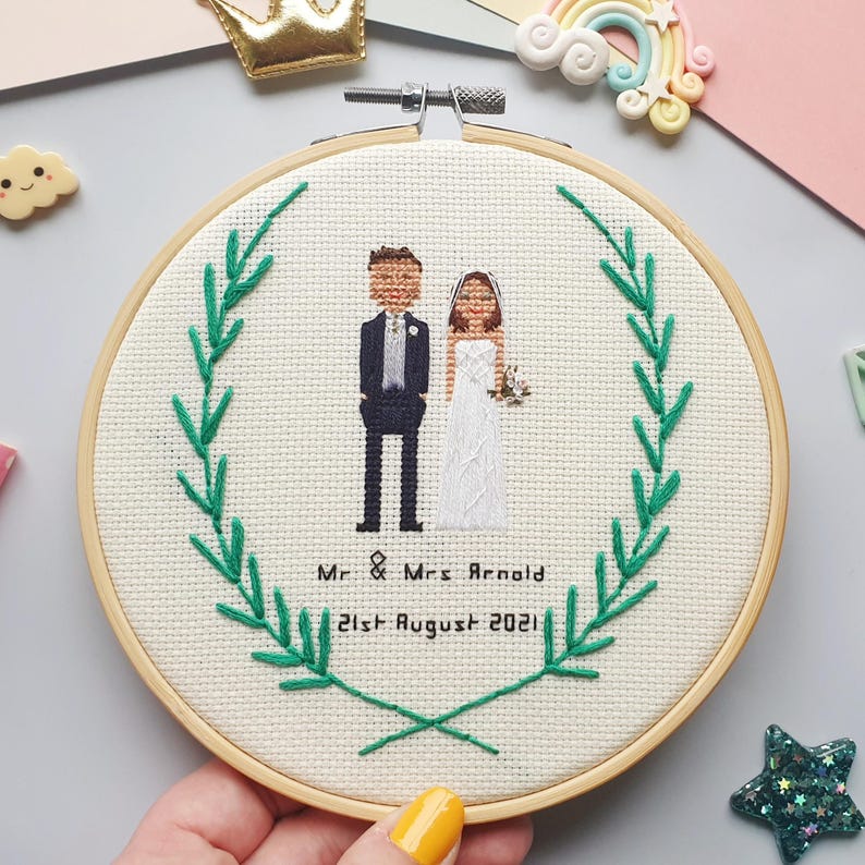 Puede incluir: Un bordado de punto de cruz circular en un aro de madera, que representa a una pareja de bodas dentro de una corona de hojas verdes. El texto "Mr & Mrs Arnold 21st August 2021" est&aacute; bordado debajo. El fondo es de tela beige claro.