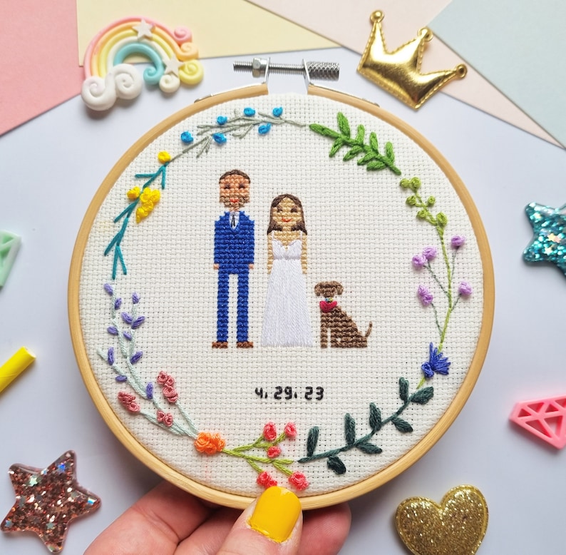 Puede incluir: Un bastidor de bordado de punto de cruz con una pareja, un perro y una corona floral. La pareja viste ropa de boda, con la fecha "4.29.23" bordada debajo. El bastidor es sostenido por una mano con esmalte de u&ntilde;as amarillo.