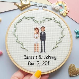 May include: A wooden embroidery hoop with two stitched figures of a bride and groom, standing under heart shaped greenery with white flowers and the text says "Genesis & Johnny Dec 2, 2011".