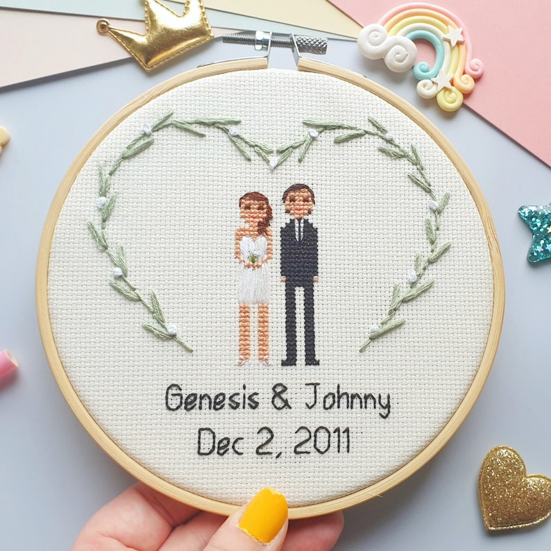 May include: A wooden embroidery hoop with two stitched figures of a bride and groom, standing under heart shaped greenery with white flowers and the text says "Genesis & Johnny Dec 2, 2011".