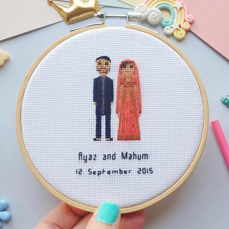 Puede incluir: Bordado de punto de cruz en un aro de madera. El dise&ntilde;o presenta una pareja con vestimenta tradicional, con los nombres "Ayaz y Mahum" y la fecha "12 de septiembre de 2015" bordados debajo. El hombre viste de azul oscuro, la mujer un vestido rojo.