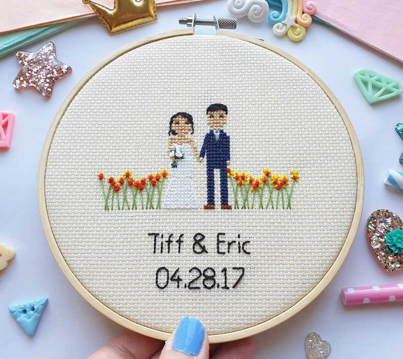 Puede incluir: Un bastidor de bordado de punto de cruz con una novia y un novio con un borde floral. Los nombres "Tiff & Eric" y la fecha "04.28.17" est&aacute;n bordados debajo. El bastidor es de madera y el fondo es de tela beige claro.