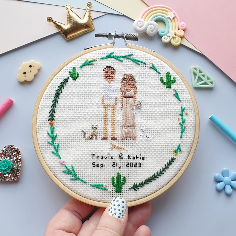 Puede incluir: Un bastidor de bordado de punto de cruz con una pareja y dos gatos, rodeado de un dise&ntilde;o floral y de cactus verdes. El texto "Travis & Katie Sep. 21, 2023" est&aacute; bordado debajo. El bastidor es sostenido por una mano con un dise&ntilde;o de u&ntilde;as de lunares.
