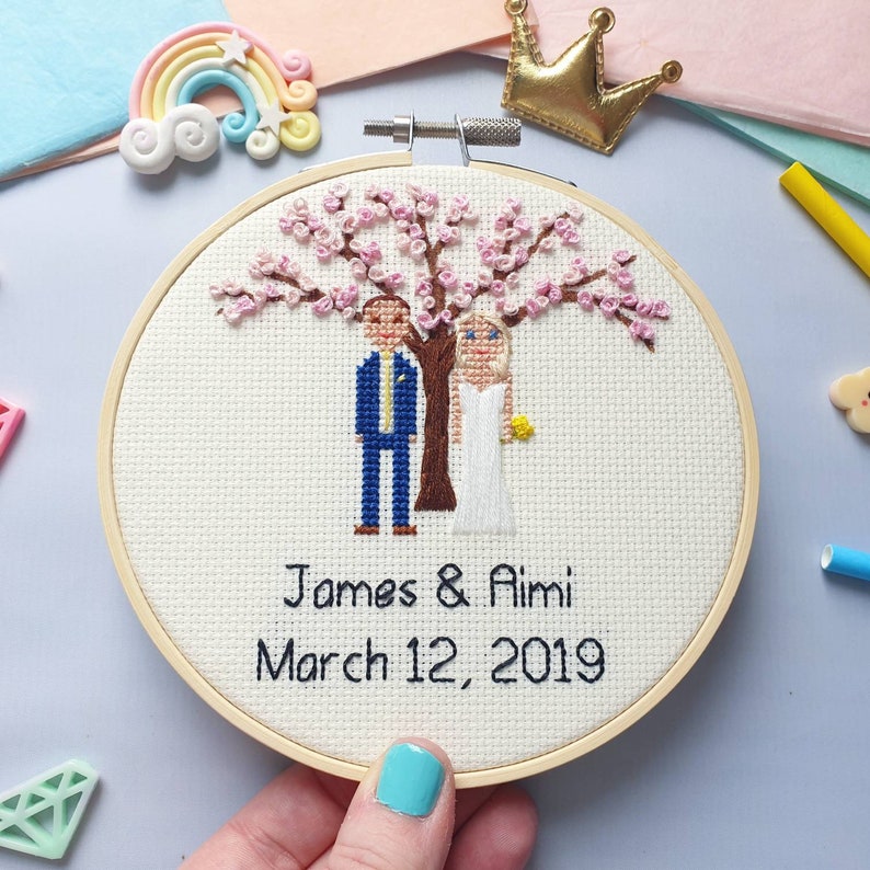 Puede incluir: Un bordado de punto de cruz en un aro de madera presenta a una pareja bajo un &aacute;rbol en flor. El hombre lleva un traje azul y la mujer un vestido blanco. Los nombres "James & Aimi" y la fecha "12 de marzo de 2019" est&aacute;n bordados debajo.